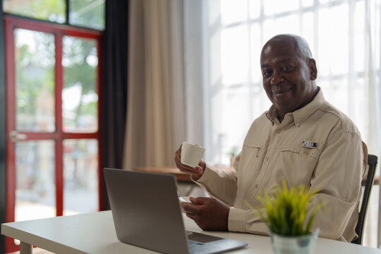Happy african american businessman drinking coffee and using laptop while working from home office, smiling mature man enjoying break time with cup of coffee and using computer - Powered by Adobe