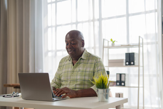Smiling african american mature male doctor wearing green shirt sitting at desk and working with laptop, stethoscope on the table, bright modern office interior, large window