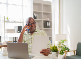 Fototapeta premium Focused senior businessman is multitasking, talking on his phone while reviewing a document at his desk in a bright, modern office setting, indicating a busy and productive workday
