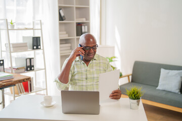Smiling, confident senior African American businessman sitting at his home office desk, holding documents while talking on a mobile phone and using a laptop
