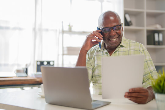 Smiling black senior manager working from a bright home office, engaging in conversation on a mobile phone while reviewing documents on a laptop, enjoying a fulfilling workday