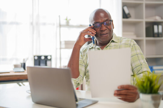 Smiling mature businessman wearing glasses, engaged in a phone conversation while reviewing a document, sitting at a desk with a laptop in a modern office environment