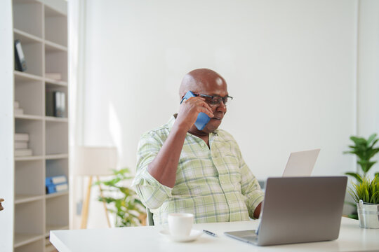 Concentrated senior businessman engaging in a phone conversation with a client while working on a laptop and reviewing documents in a well-equipped home office environment