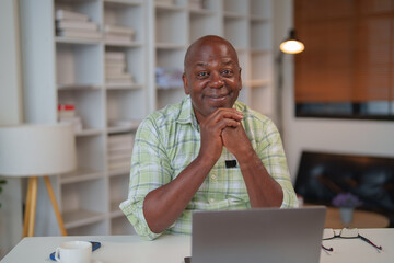 Happy african american businessman is sitting at desk and working remotely from home office using laptop, he is smiling and looking confident