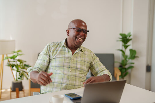 Cheerful african american man wearing glasses is sitting at his desk, using his laptop and laughing while having a video call with his family or friends