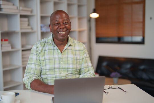 Happy African American mature male freelancer working remotely, sitting comfortably at a desk in a cozy home office, with a bookshelf filled with books in the background