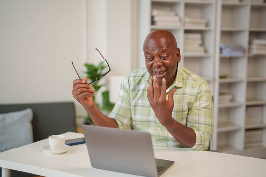 Surprised senior african american man in casual clothes is holding his eyeglasses and having a video call on his laptop while sitting at his desk in his modern home office - Powered by Adobe