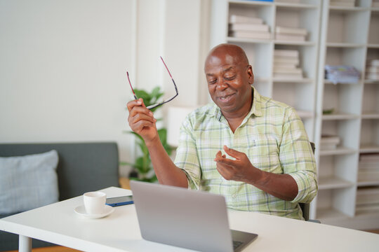 Smiling mature African American teacher holding eyeglasses and gesturing while explaining concepts during an engaging online lesson on a laptop at home, creating an interactive learning environment