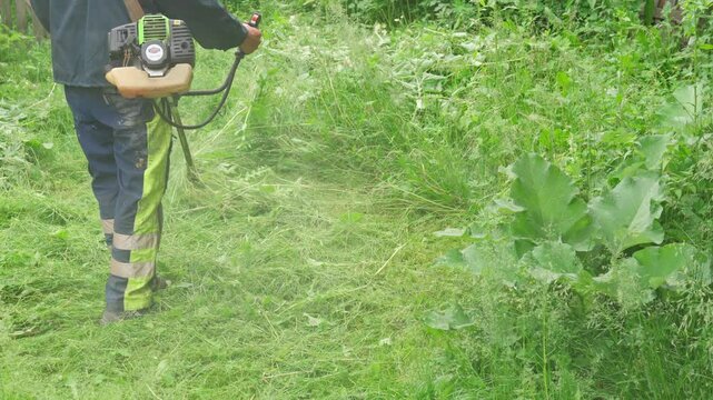 Worker mowing grass with a trimmer. Man cutting green grass using brush cutter. Lawn mowing service. Gardener using lawnmower outdoors.