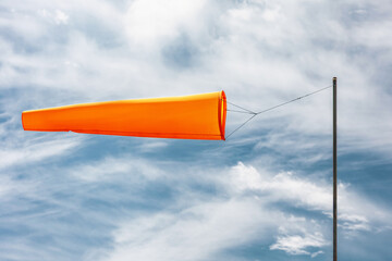 Orange windsock is attached to a pole and showing the wind direction and speed on a cloudy sky