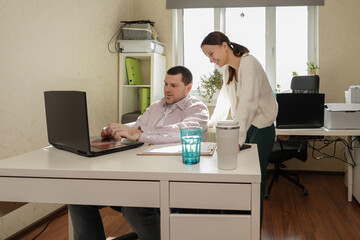 Obraz premium Casual work environment where a man is seated in an ergonomic chair typing while a standing female colleague offers feedback, both framed by office supplies