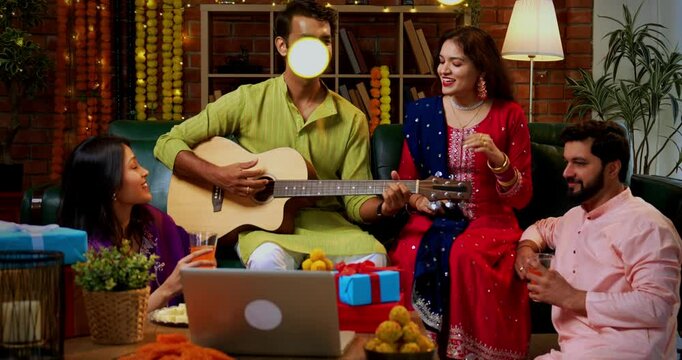 Friends singing during Diwali celebration, Indian Asian young married couple hosting friends with guitar and music in traditional festive wear enjoying Deepavali night at decorated home with sweets