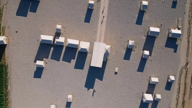 Topdown video of scattered modular shelters casting long shadows on a gravel-covered surface under strong sun

