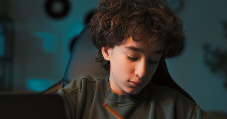 A boy works on test preparation by rewriting notes with a pencil, studying online from an e-book while attending a remote schooling session from home.