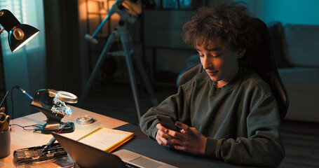A smart boy attends remote lessons on his laptop while searching for test answers on his phone. He reviews research, chats on social media, and studies school content with focus.