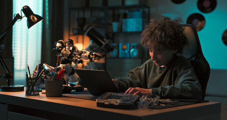 A young talented boy sits in a dark room, programming a robot on his laptop. He adds new features and high-fives the toy with pride, showing love for robotics and innovation.