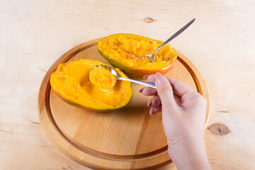 Side view of woman hand holding the spoon with mango pulp and two halves of mango on wooden background