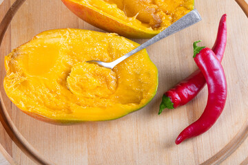 Top view of two halves of mango, two red hot chili peppers fnd spoon on wooden background