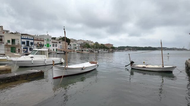 White lla&uuml;t moored in the calm waters of portocolom, mallorca, spain, with colorful houses in the background on a cloudy day