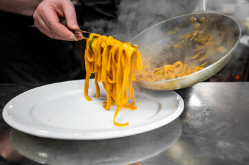 A chef serves freshly cooked fettuccine pasta from a steaming pan onto a white plate. The dish is vibrant, seasoned with herbs, and presented in a professional culinary setting