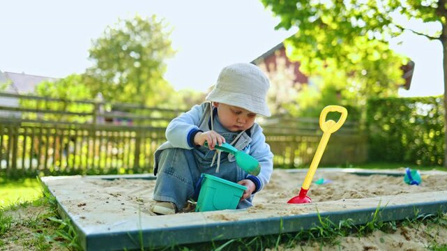 Two-year-old boy in a Panama hat on his head plays in a sandbox with sand on a playground. Toddler in stylish casual clothes digs sand while sitting in a sandbox on the street. Child development.