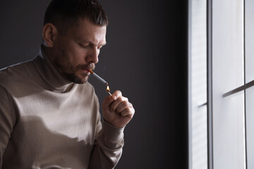 Man lighting cigarette with match near window indoors, space for text