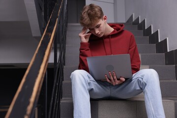 Cyber bullying. Upset teenage boy with laptop on stairs indoors