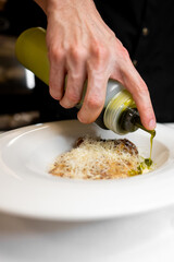 A close-up of a hand pouring vibrant green sauce from a squeeze bottle onto a freshly prepared pasta dish, garnished with grated cheese. A culinary scene capturing food presentation