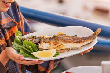 Smiling waitress holding a plate of grilled sea bass with lemon and arugula, in a restaurant by the...