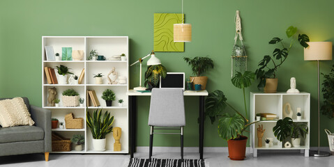 Stylish workplace with laptop, shelving unit and different houseplants near green wall at home