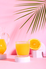 Decorative podium with glass of fresh orange juice and palm leaf on pink background
