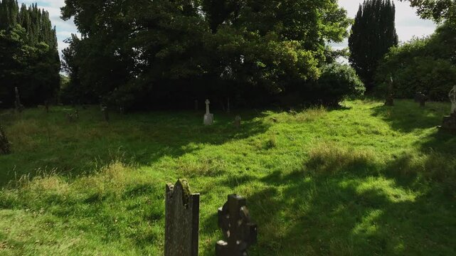 Old Donagh Graveyard, County Monaghan, Ireland, September 2022. Drone pullback over grass as sunlight filters through trees illuminating tomb and headstones weathered from age.