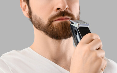 Handsome young man shaving his beard with electric shaver on grey background, closeup