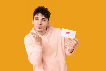 Handsome young man holding envelope with lipstick mark and blowing kiss on yellow background