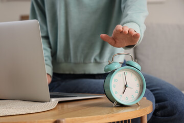Woman with laptop turning off alarm clock on table at home, closeup