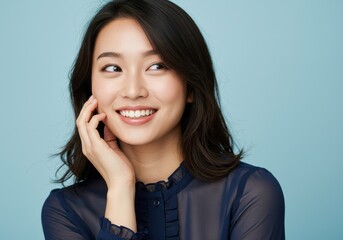 Fototapeta premium Smiling young Asian woman with hand near face against light blue background