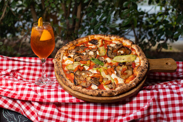 Wood-fired vegetable pizza and orange cocktail on checkered picnic cloth.
