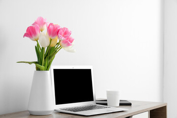 Stylish workspace with modern laptop and beautiful flowers near white wall