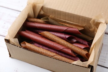 Tasty fruit leather rolls in cardboard box on white wooden table, closeup