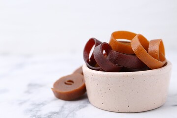 Tasty fruit leather rolls in bowl on white marble table, closeup. Space for text