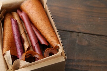 Delicious fruit leather rolls in cardboard box on wooden table, closeup. Space for text