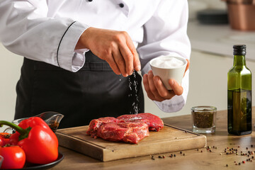 Male chef salting raw meat at table in kitchen, closeup