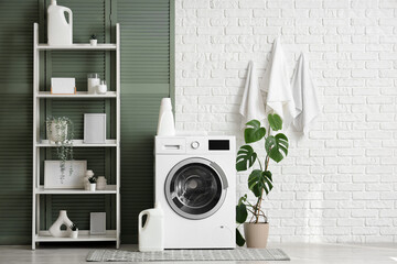 Modern washing machine with shelving unit, houseplant and detergents near white brick wall in...