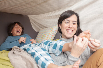Smiling mother holding son's foot on her shoulder while he laughs in sofa fort