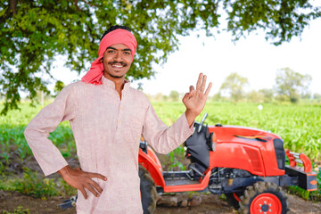 Happy young indian farmer standing with new tractor at agriculture field