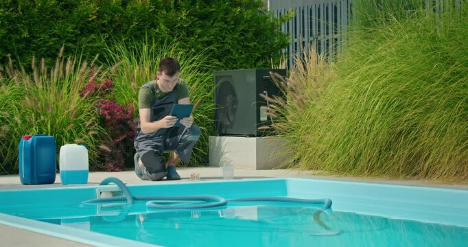 Pool technician kneels beside a backyard swimming pool, checking chemical levels using a digital tablet with containers of pool chemicals nearby.