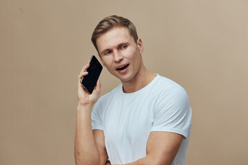 young smiling man talking on mobile phone wearing white t-shirt standing isolated on beige...