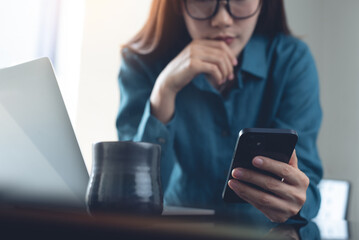 Business woman using mobile phone during working on laptop computer at office. Young female entrepreneur using smartphone looking at phone screen, thinking of problem solution, working at home office
