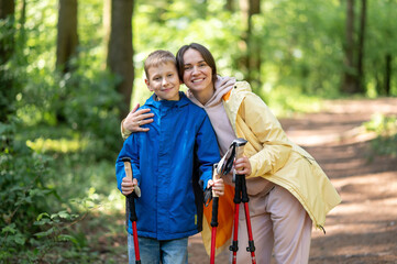 Mother hugging her young son in a sunny forest during a Nordic walking break, sharing a loving and...