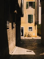 Sunlight illuminating narrow cobblestone alley with yellow wall and black bicycle in Rovinj, Croatia, creating strong shadows on stone pavement. Yellow wall framing vintage bicycle in Rovinj alleyway